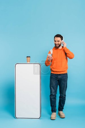 full length of bearded student in wireless headphones standing with smartphone near phone mock-up and paper cup on blue background