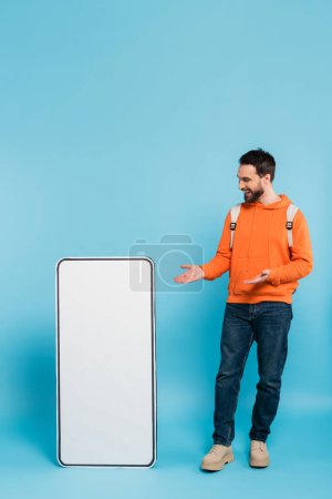 Photo for Full length of happy bearded student pointing at big template of mobile phone on blue background - Royalty Free Image