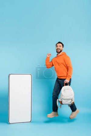 Foto de Full length of cheerful bearded student with backpack showing win gesture while levitating near phone template on blue background - Imagen libre de derechos