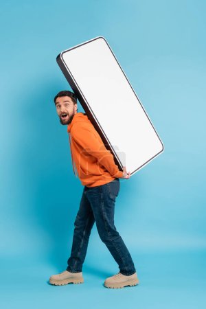 full length of amazed man in orange hoodie and jeans carrying carton phone mock-up on blue background