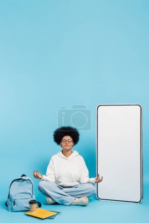 african american student in eyeglasses meditating in lotus pose near backpack and cardboard phone template on blue background