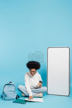 african american student with paper cup looking in notebook while sitting near backpack and white phone mock-up on blue 