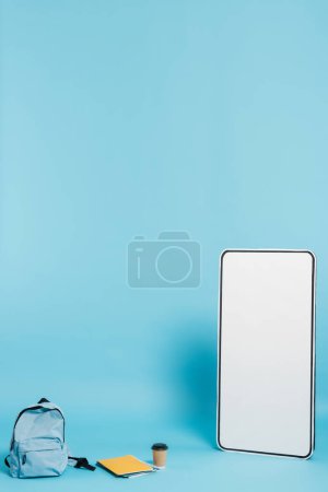 Photo for Vertical view of white phone template near backpack and notebooks with paper cup on blue background - Royalty Free Image
