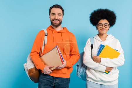 Photo for Cheerful multiethnic students in hoodies with copybooks and backpacks looking at camera isolated on blue - Royalty Free Image