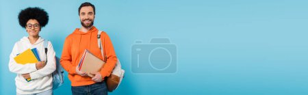 Photo for Positive interracial students with backpacks and notebooks smiling at camera isolated on blue, banner - Royalty Free Image