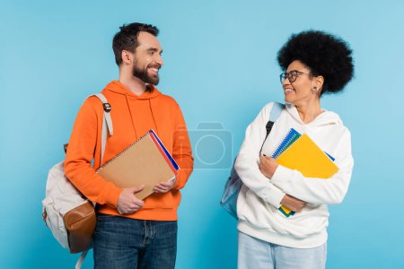 Photo for Happy and stylish interracial students with backpacks and notebooks looking at each other isolated on blue - Royalty Free Image