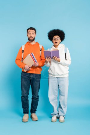 Photo for Full length of shocked multiethnic students with backpacks and notebooks looking at camera on blue - Royalty Free Image