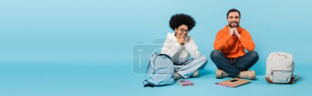 cheerful interracial students sitting with crossed legs near backpacks and smartphones  on blue background, banner
