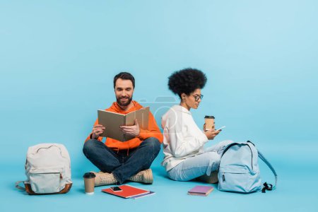 Photo for Bearded student looking in notebook near african american girlfriend sitting with smartphone on blue background - Royalty Free Image