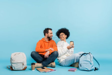 Photo for Happy multiethnic students sitting near backpacks and copybooks and smiling at each other on blue background - Royalty Free Image