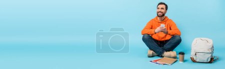 Photo for Full length of happy bearded student with smartphone sitting near backpack and paper cup with notebooks on blue background, banner - Royalty Free Image