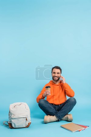 Photo for Smiling student sitting with coffee to go and talking on smartphone near backpack and copybooks on blue - Royalty Free Image