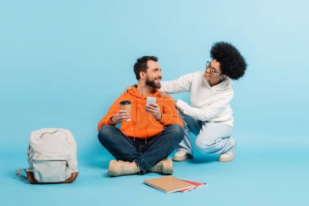 african american woman touching shoulders of bearded student sitting with smartphone and paper cup on blue 