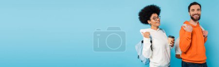 Photo for Happy interracial students with backpacks and coffee to go pointing with thumbs and looking at camera isolated on blue, banner - Royalty Free Image