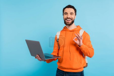 happy bearded student holding laptop and showing okay sign isolated on blue