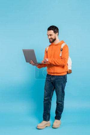 full length of happy student in orange hoodie and jeans using laptop on blue 