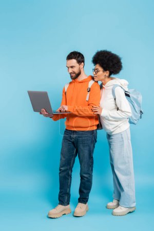 full length of bearded student using laptop near african american girlfriend on blue 