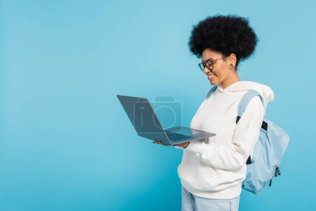 cheerful african american student with backpack looking at laptop isolated on blue