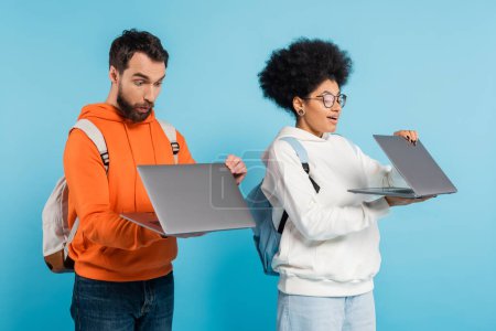 Photo for Amazed interracial students with backpacks looking at laptops isolated on blue - Royalty Free Image