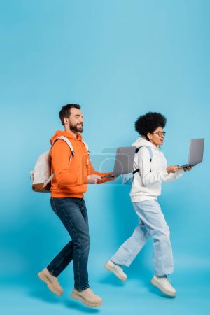 full length of stylish multiethnic students with backpacks and laptops levitating on blue background