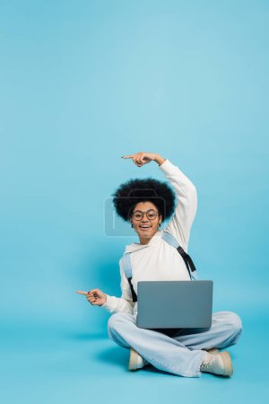 full length of smiling african american student sitting with laptop and pointing with fingers on blue background