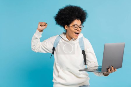 excited african american student in eyeglasses looking at laptop and showing win gesture isolated on blue