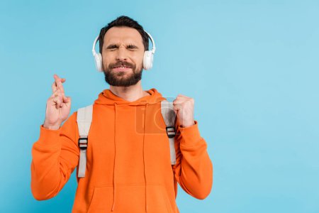 positive bearded student in wireless headphones crossing fingers while standing with closed eyes isolated on blue