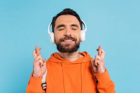 Photo for Positive student in wireless headphones holding crossed fingers while standing with closed eyes isolated on blue - Royalty Free Image