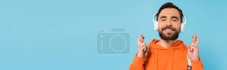 Photo for Smiling student in wireless headphones crossing fingers with closed eyes isolated on blue, banner - Royalty Free Image