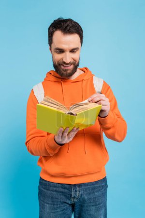 Téléchargez les photos : Smiling bearded student in orange hoodie reading book isolated on blue - en image libre de droit