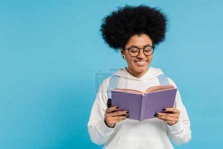 curly and stylish african american student in eyeglasses smiling while reading book isolated on blue