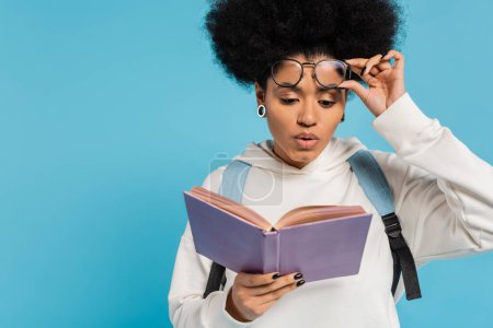 impressed african american student holding eyeglasses while reading book isolated on blue