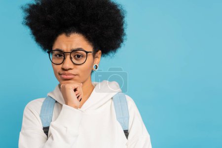 upset and thoughtful african american student in eyeglasses holding hand near chin and looking at camera isolated on blue