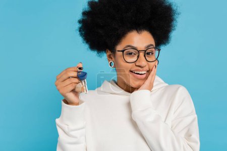Photo for Happy african american woman in stylish eyeglasses and white hoodie touching face while holding key isolated on blue - Royalty Free Image