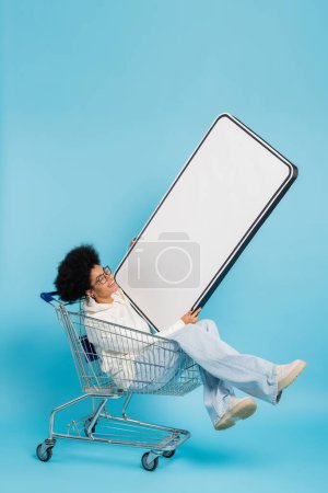 full length of excited african american woman sitting in shopping cart with big phone template on blue background