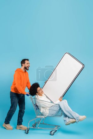 Photo for Bearded man laughing near shopping cart with african american woman and mobile phone template on blue background - Royalty Free Image