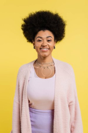 cheerful african american woman in soft cardigan and silver necklaces looking at camera isolated on yellow