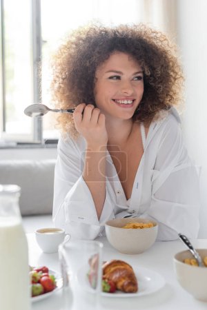 cheerful young woman holding spoon while having breakfast in kitchen 