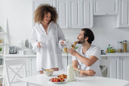 curly woman holding cups of coffee near bearded boyfriend during breakfast  