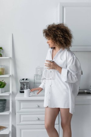 curly young woman in white shirt standing with cup of coffee in kitchen 