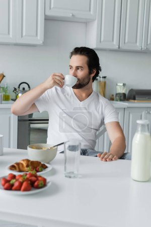 bearded man drinking coffee near tasty breakfast on table in kitchen 