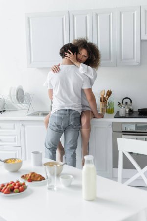 back view of man in jeans seducing curly woman sitting on kitchen worktop 
