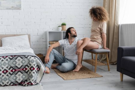 bearded man sitting on carpet and looking at curly girlfriend on bed bench 