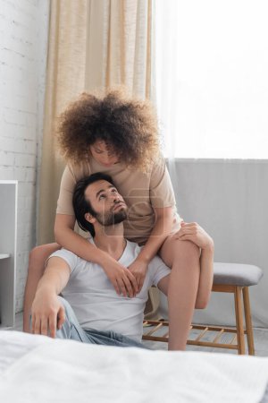 curly woman sitting on bed bench hugging bearded boyfriend in bedroom 