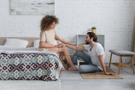 full length of barefoot woman sitting on bed and touching hand of bearded boyfriend in bedroom 