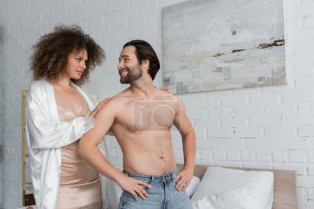 sexy woman in silk robe looking at shirtless boyfriend in jeans posing with hands on hips 
