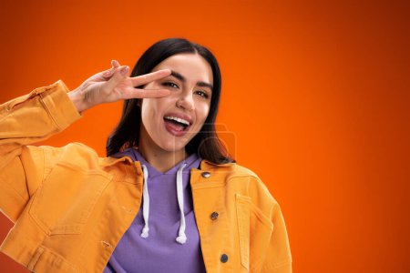 Cheerful young woman showing peace sign at camera isolated on orange