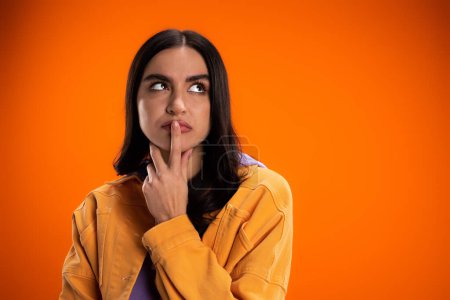 Pensive brunette woman touching lips and looking away isolated on orange