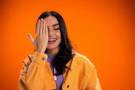 Photo for Smiling brunette woman covering face isolated on orange - Royalty Free Image