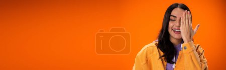 Foto de Cheerful brunette woman covering face isolated on orange, banner - Imagen libre de derechos